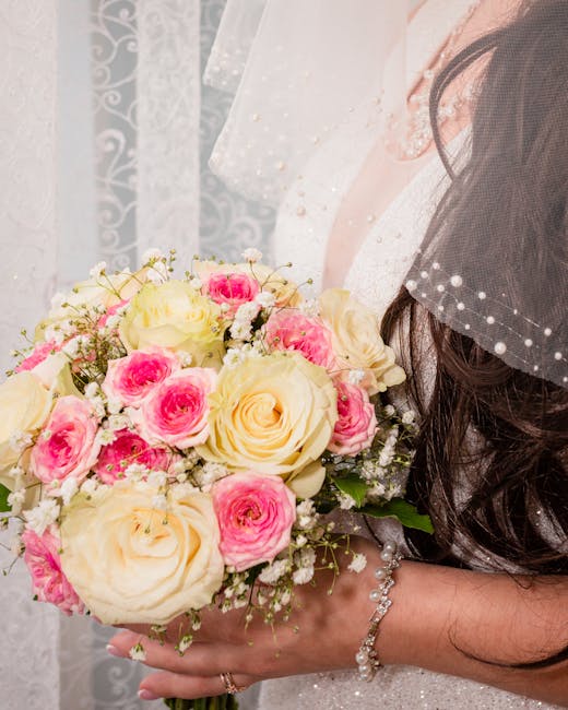 A bride holds a floral bouquet consisting of large ivory roses and smaller pink and white spray roses, accented with delicate baby's breath and green foliage. The bouquet is wrapped with a simple whit
