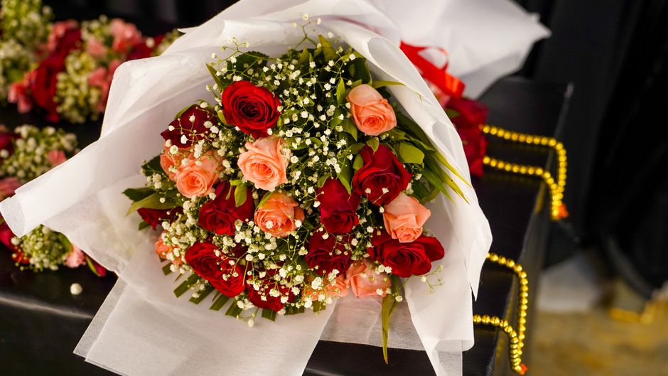 A vibrant bouquet of fresh roses arranged with pink and red blooms, accented by delicate white baby's breath and lush green foliage. The flowers are wrapped in elegant white tissue paper, with a hint 