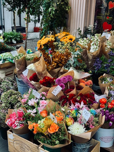 An outdoor display of fresh flower bouquets arranged in various containers, including metal buckets and cardboard wrapping, featuring a vibrant mix of flowers such as sunflowers, roses, tulips, chrysa