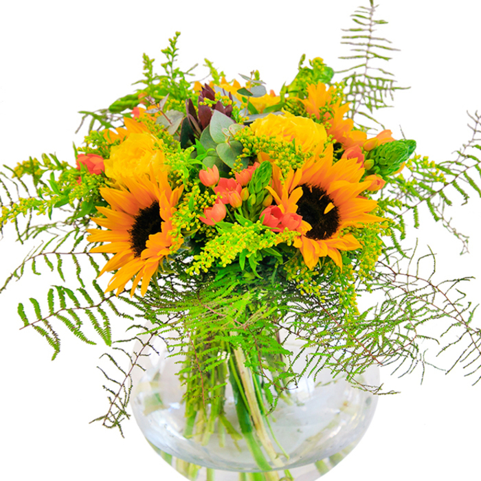 The immediate impression of this bouquet is one of brilliant sun-drenched cheer, much like the vibrant scenes of Dartford's open spaces. Sunflowers, their faces boldly turned outwards, radiate yellow and orange hues alongside classic garden roses in a buttery palette, each rose curling in tight, elegant spirals. Scarlet Hypericum berries punctuate the greenery, sparkling exuberantly and providing a lively contrast to the softer petals. A tumble of feathery ferns and sprigs of solidago with their tiny yellow blossoms create a cascading frame, reminiscent of the wild edges found along the Darent River. Bluish-green eucalyptus and slender spikes of green carry the eye through every curve and detail. The spherical glass vase in which the arrangement sits reflects Dartford's blend of tradition and modernity, showcasing the bouquet's architectural grace and the freshness of every cut stem. The subtle perfume hints at meadow walks and garden celebrations. Suitable for special occasions-birthdays, anniversaries, or simply adding joy to an ordinary day-this arrangement encapsulates the artistry and heartfelt care synonymous with Florist Dartford.