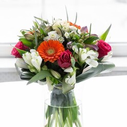 This splendid bouquet pays homage to Dartford's vibrant sense of community and natural beauty, as if gathered from the flower fields just beyond Darenth. At its core, orange and peach gerberas unfurl their plush petals in a welcoming embrace, glowing like the sunrise over the town centre. Deep magenta roses nestle beside them, rich and regal, their compact petals carrying an aura of romance and refined artistry. Accents of ivory alstroemeria and fluffy white spray roses stretch between the bolder tones, brightening the entire composition with an airy, gentle touch that brings to mind fresh blooms at the entrance of Market Street. Touches of soft pink and tiny filler flowers create layers of abundance, their hues peeking out to foster visual intrigue. The base is well-supported by a flourish of glossy, deep green leaves, including wide, curved aspidistra leaves that form a graceful, protective rim-mirroring Dartford's leafy avenues and welcoming green parks. Fine, pointed greenery is deftly woven throughout for texture and contrast. Presented in a tall, clear glass vase, the bouquet's stems are visible, lending authenticity and life. Illuminated by diffuse light, every colour and shape is celebrated without distraction. This bouquet captures the spirit of joyful gathering, appreciation, and major milestones, perfect for adding a splash of Dartford's signature brightness to any setting or occasion.